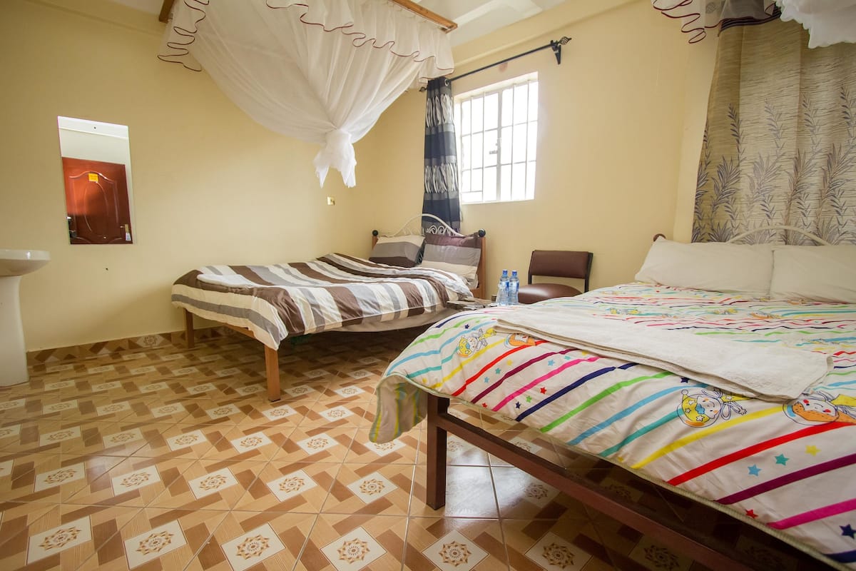A bedroom with two beds, a window with curtains, a bedside table with a water bottle, and a patterned floor.
