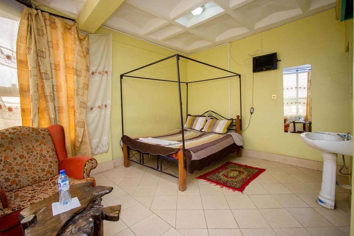 A room with a four-poster bed, a sofa, a wooden table with a water bottle, a red and gold rug, a white sink, and a television on the wall.