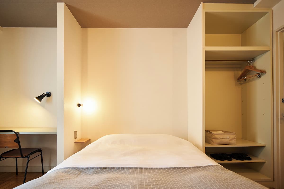A bedroom with a bed, a desk with a chair, a desk lamp, and a wardrobe with shelves.
