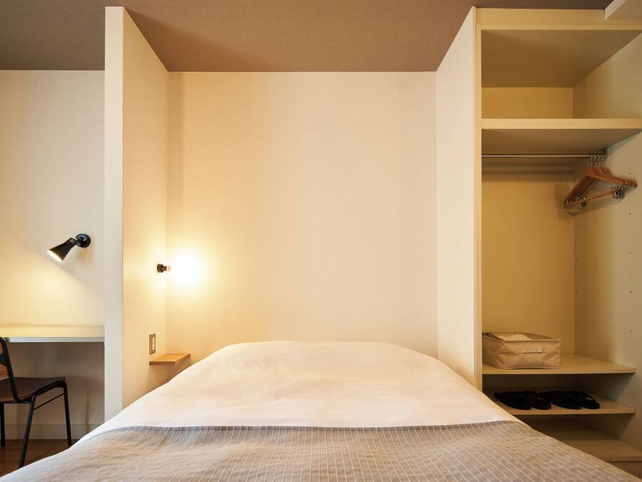 A bedroom with a bed, a desk with a chair, a desk lamp, and a wardrobe with shelves.
