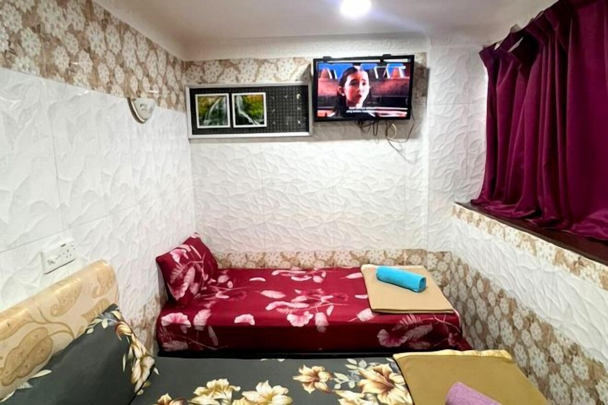 A small, furnished room with a bed, a sofa, a TV mounted on the wall, and a window with curtains.