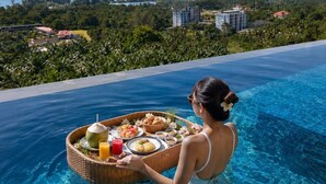 A woman in a pool holding a tray with food and drinks, with a view of buildings and mountains in the background. - Andakiri Pool Villa Panoramic Sea View (Krabi)
