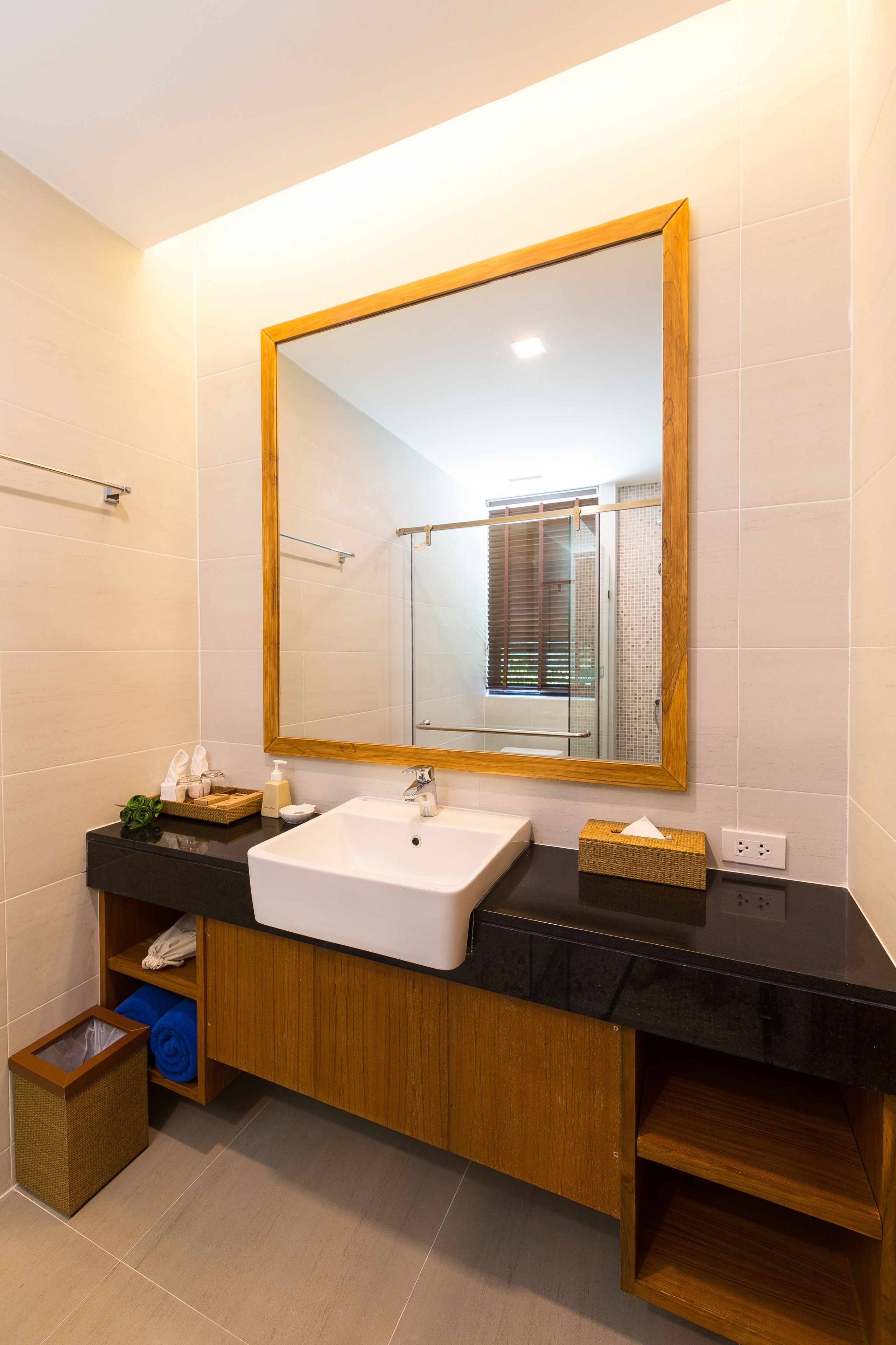 A modern bathroom with a large mirror, white sink, wooden vanity, and a small trash bin.