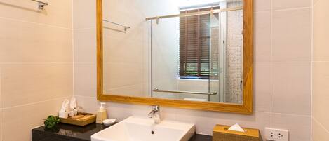 A modern bathroom with a large mirror, white sink, wooden vanity, and a small trash bin.