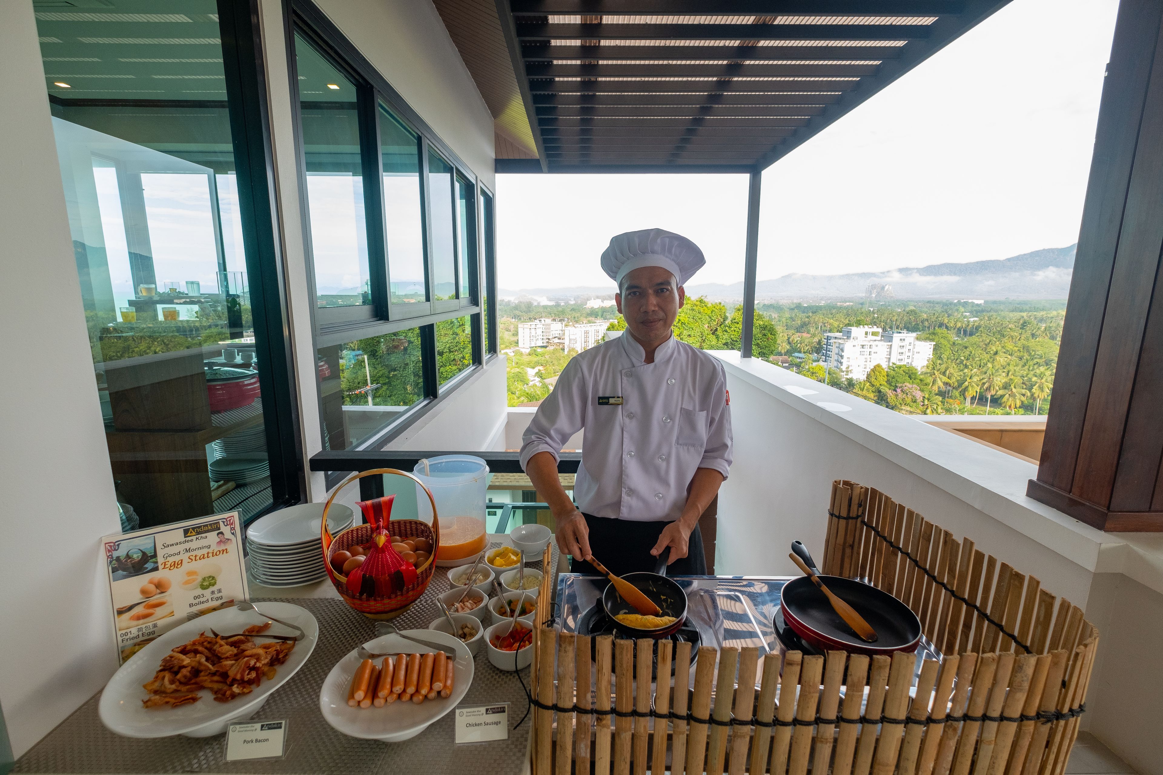Een kok bereidt eten op een balkon met uitzicht op de stad, een buffetopstelling met diverse gerechten, en een bordje dat een eilandje aangeeft.