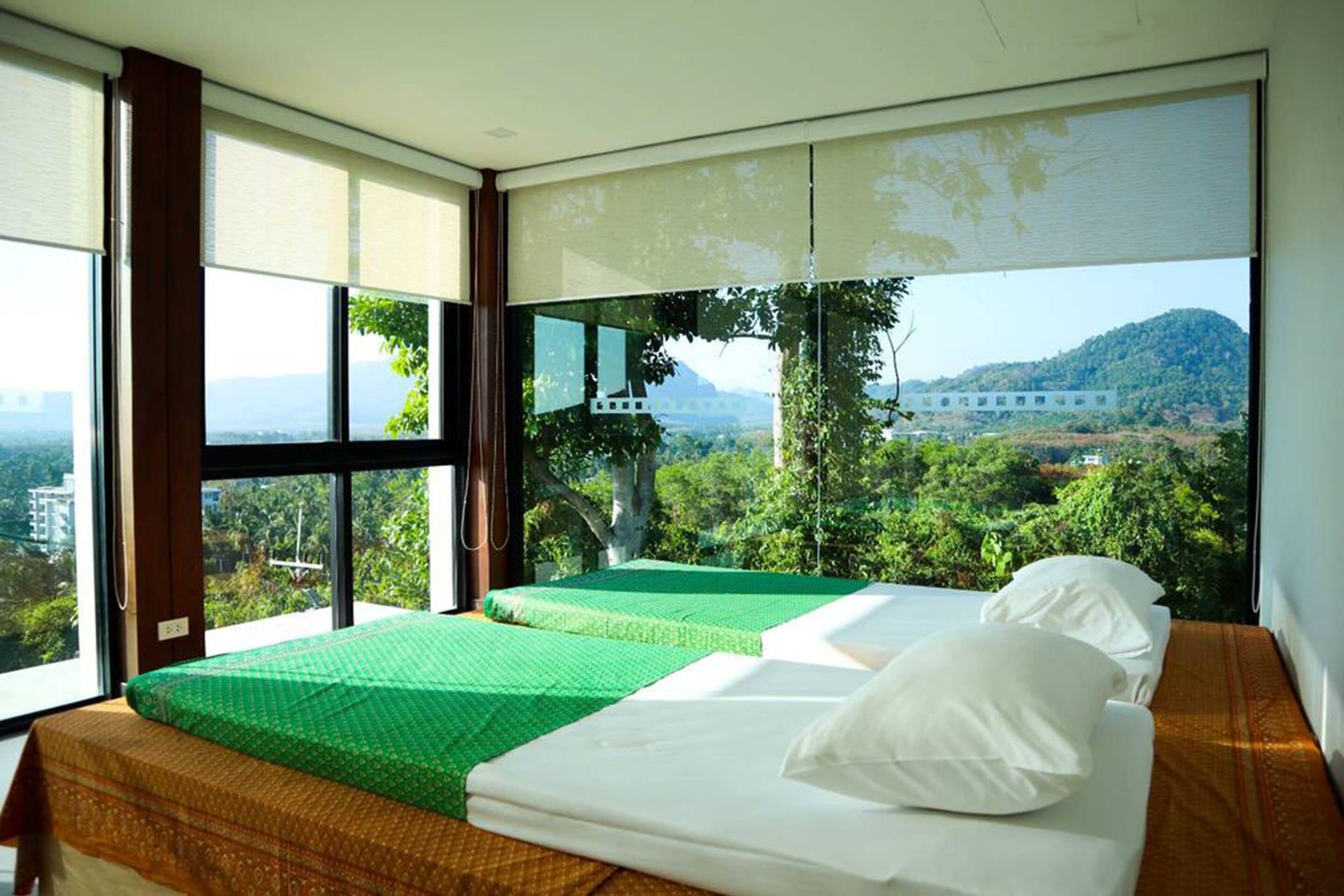 A room with two beds, a large window with a view of greenery and a distant building, and a ceiling fan.
