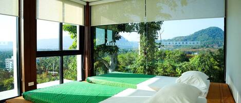 A room with two beds, a large window with a view of greenery and a distant building, and a ceiling fan.