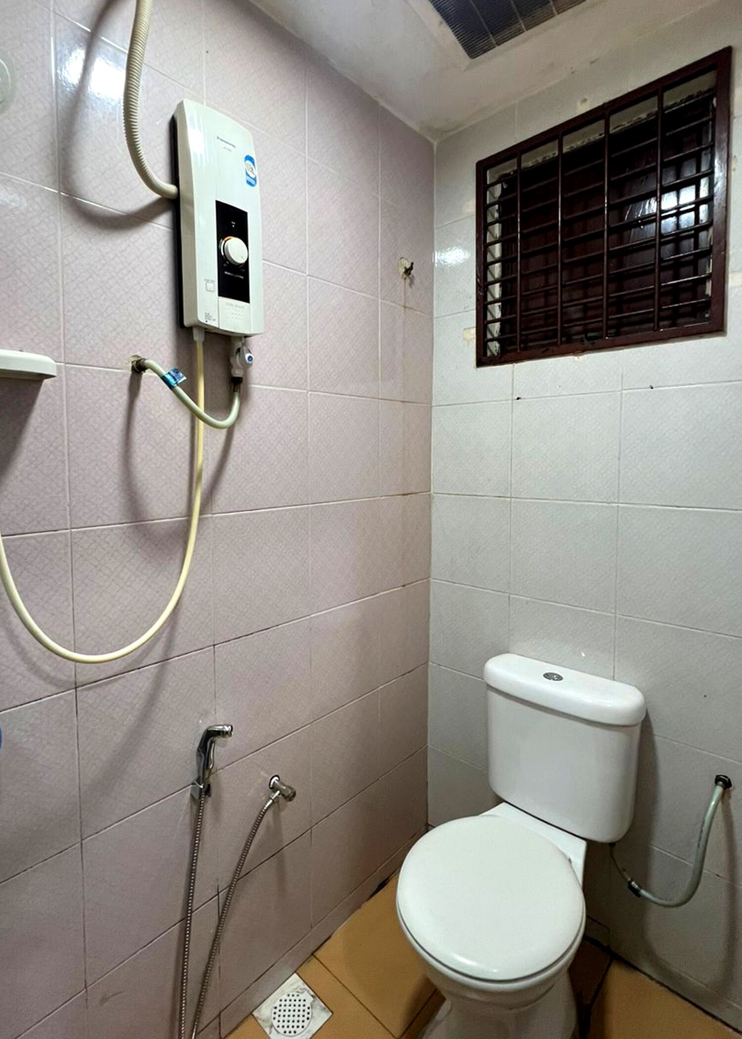 A small, tiled bathroom with a toilet, a shower, and a window with bars.