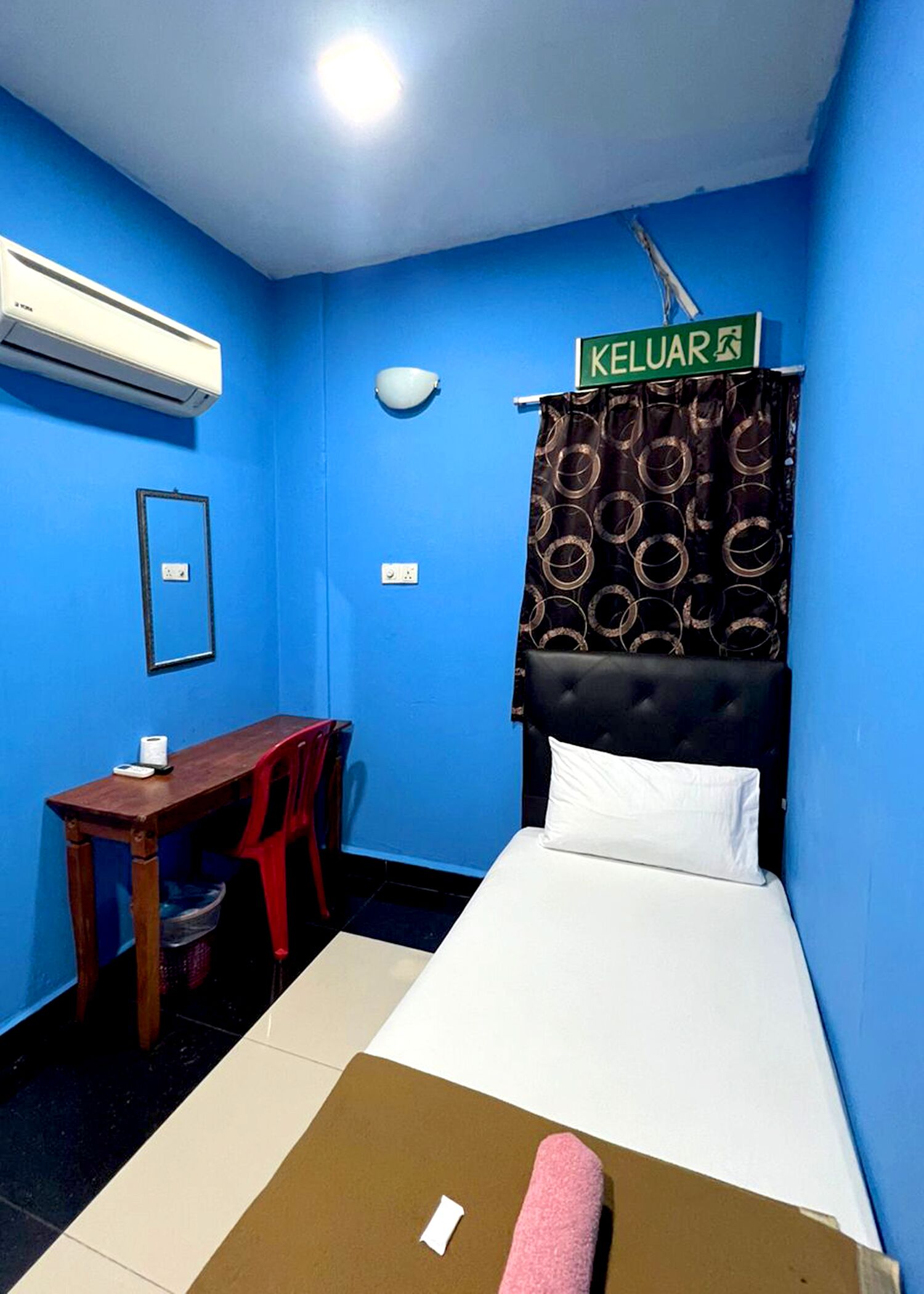 Quarto com cama, escrivaninha, cadeira e ar-condicionado, e um aviso com a inscrição 'KELUAR'.