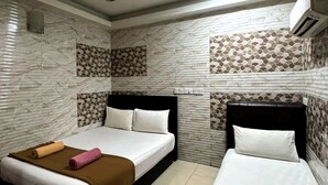 A hotel room with two beds, each with a brown bedspread and white linens. The walls have a patterned tile design, and there is a ceiling light.