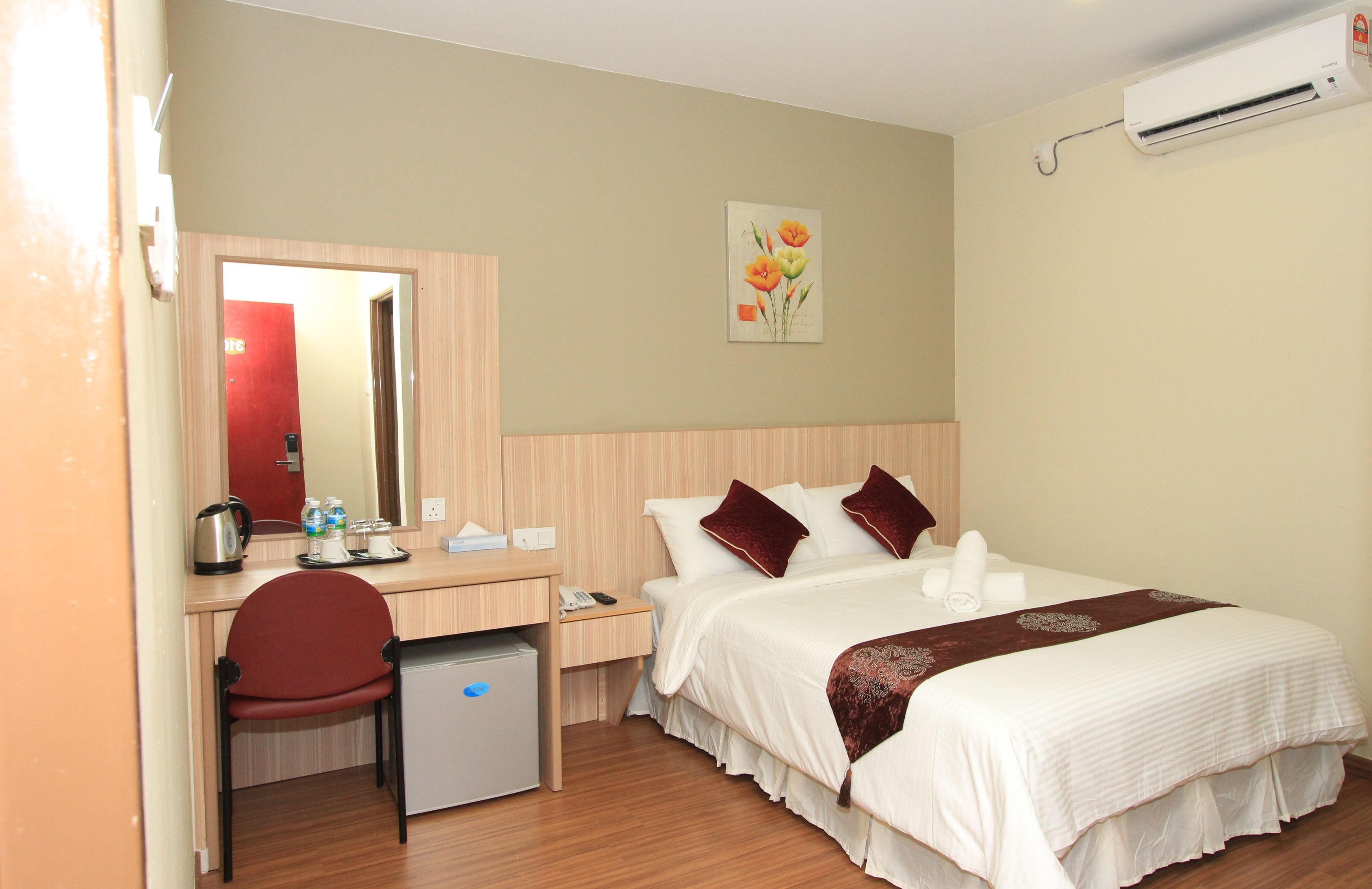 Habitación de hotel con cama, escritorio con silla, refrigerador pequeño, espejo, aire acondicionado de pared y un cuadro de flores.