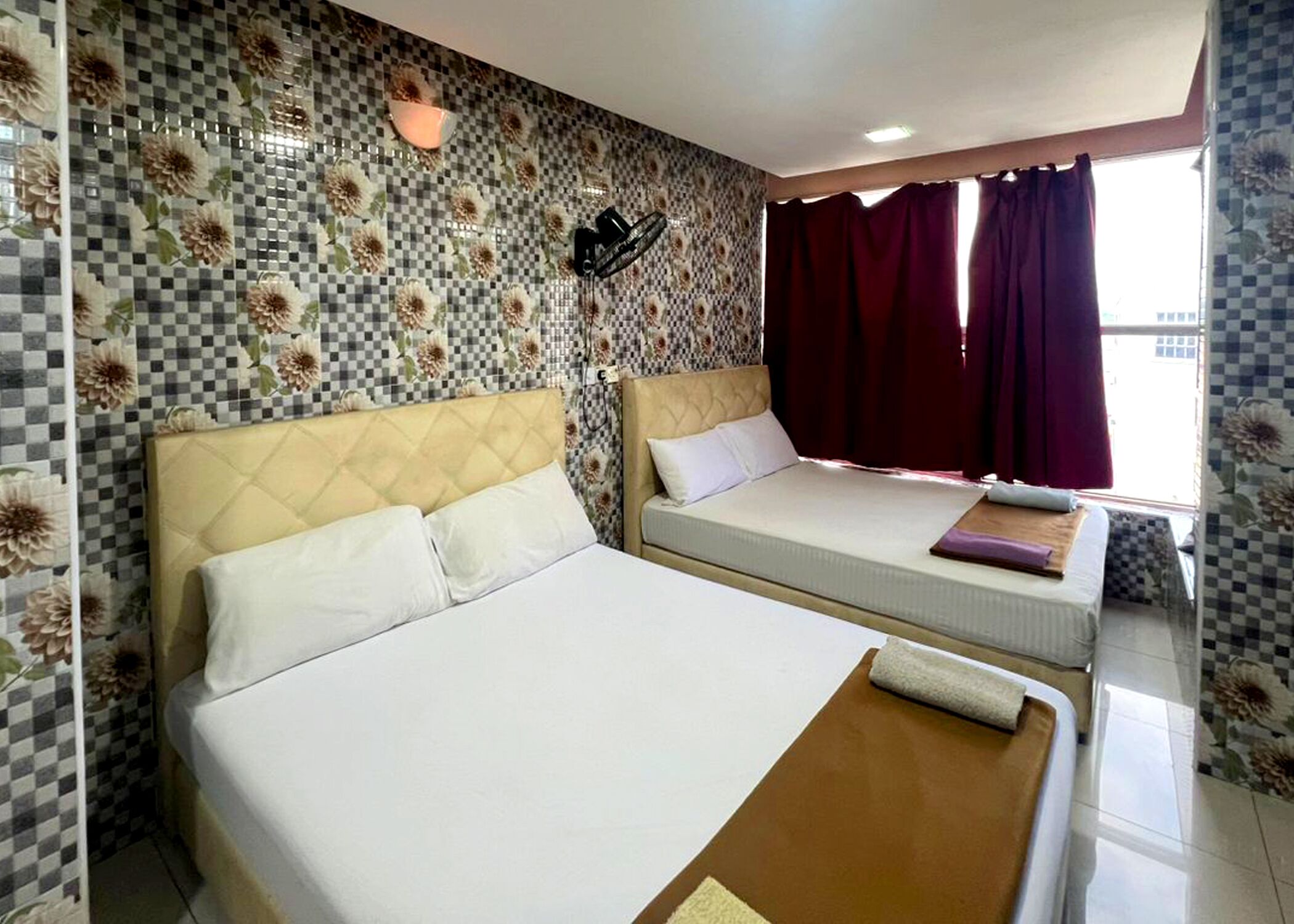 A hotel room with two beds, checkered floral wall tiles, a window with red curtains, and a wall-mounted light fixture.