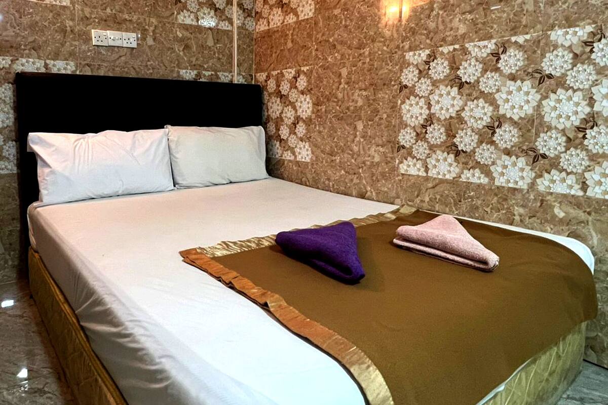 Un letto rifatto con lenzuola bianche, una coperta marrone e due asciugamani, uno viola e uno rosa, in una stanza con pavimento in marmo e pareti a motivi floreali.