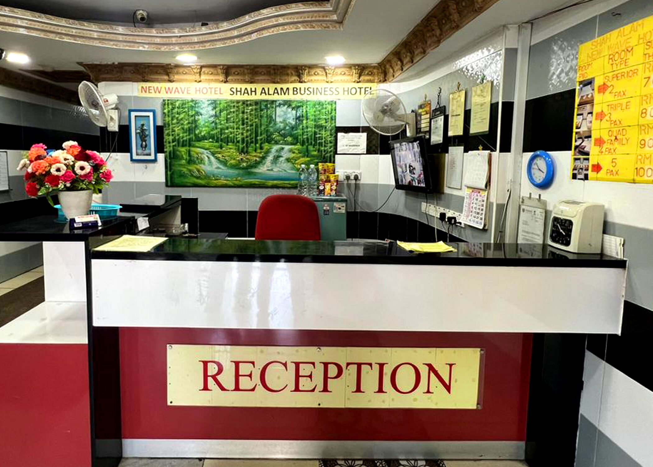 A reception area with a 'RECEPTION' sign, a large painting of a forest, a desk with a computer, a red chair, and a wall with a yellow sign listing room types and prices.