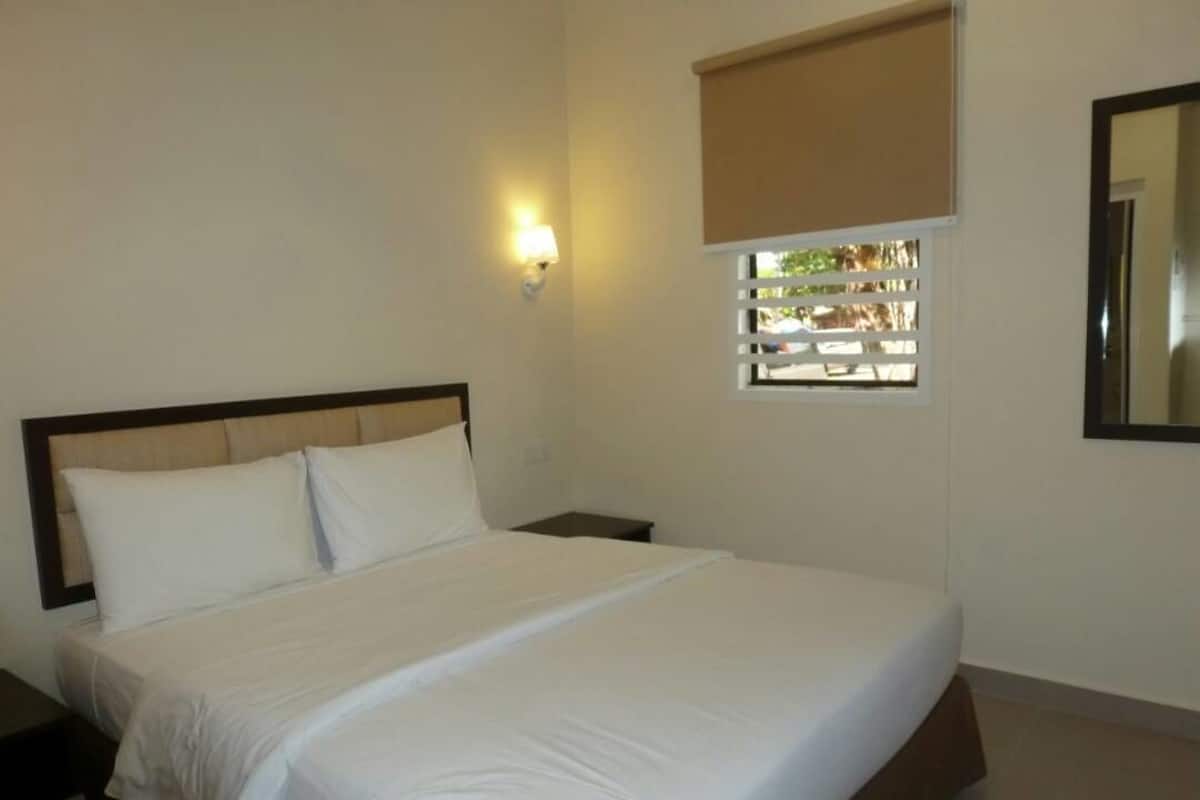 Ein Schlafzimmer mit einem großen Bett, einem Nachttisch, einer Wandlampe, einem Fenster mit Jalousien und einem Spiegel.