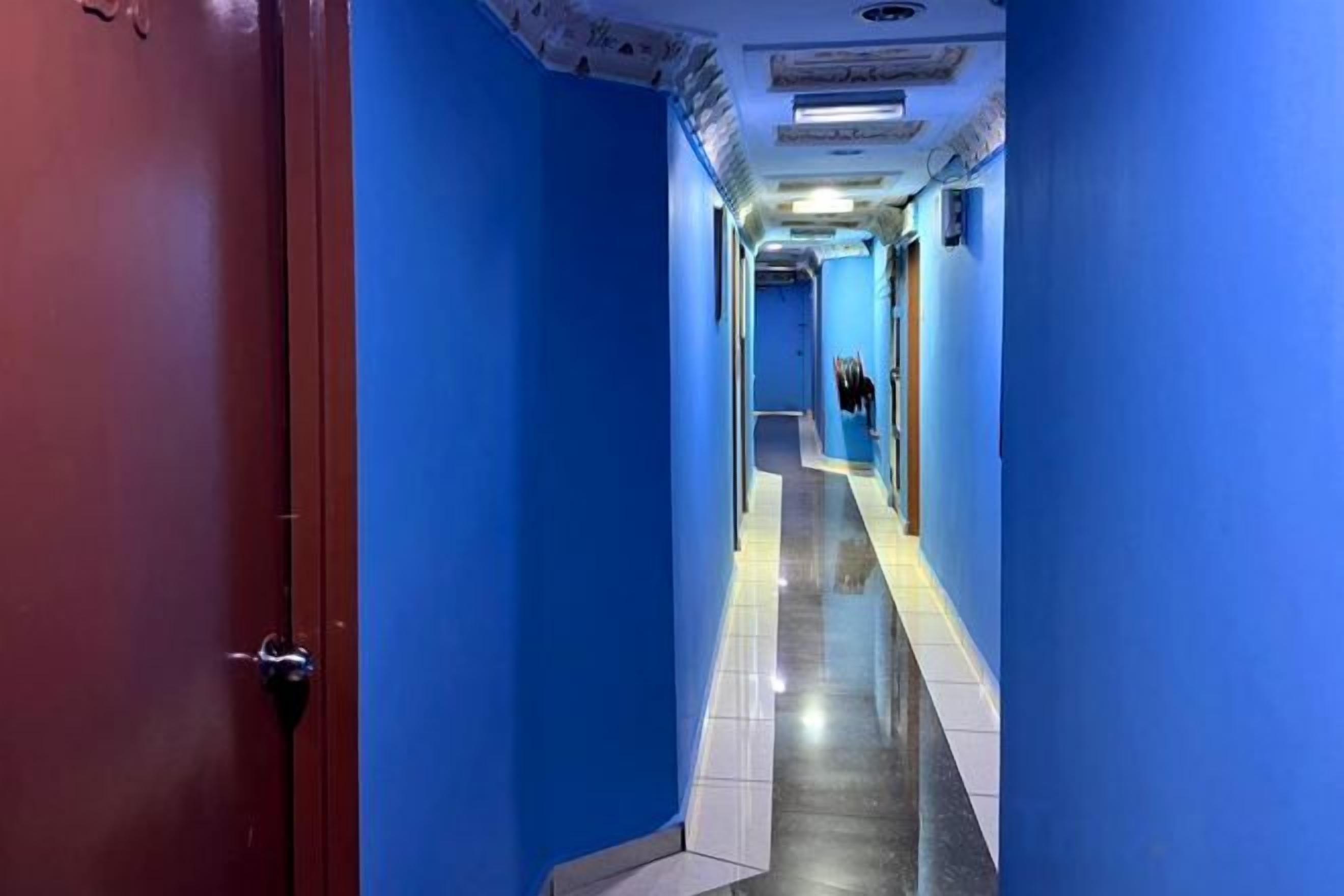 A corridor with blue walls and a red door, leading to a room with a bed and a window.