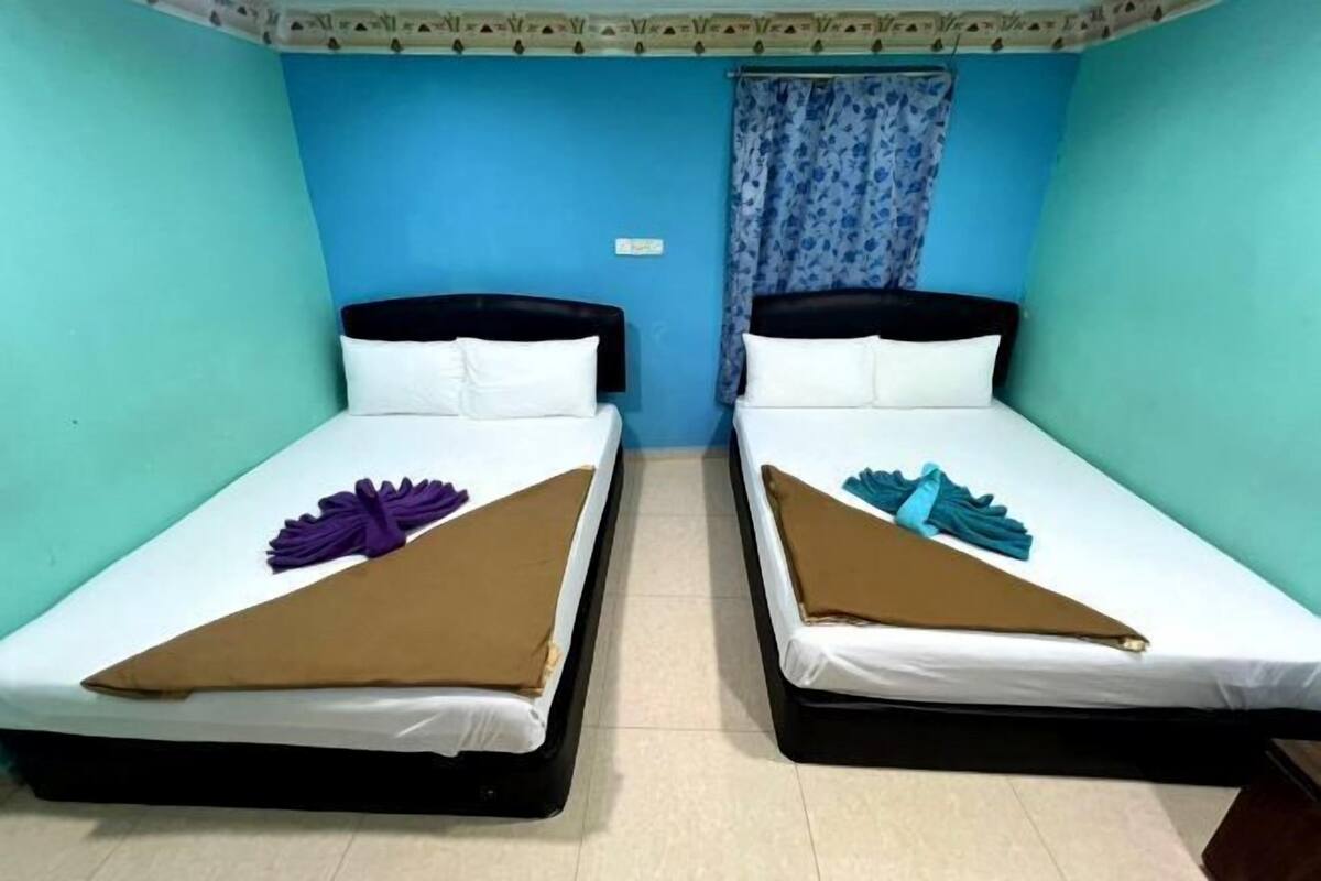 Een kamer met twee bedden, elk met een zwart hoofdbord, witte beddengoed en een bruine deken. Er hangt een gordijn met een bloemmotief aan een wand.