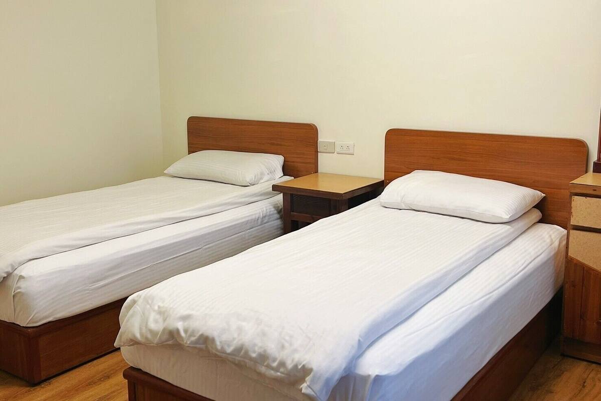 A room with two single beds, wooden headboards, a small wooden nightstand, and a light switch on the wall.