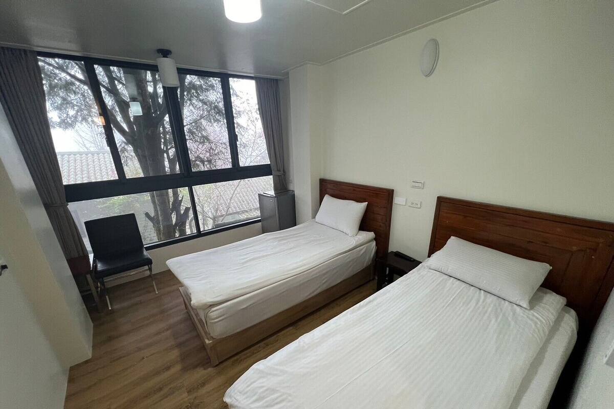 A hotel room with two beds, a desk, and a window with a view of trees.