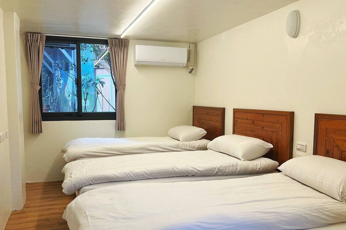 A hotel room with two beds, a window with curtains, an air conditioning unit, and a light switch on the wall.