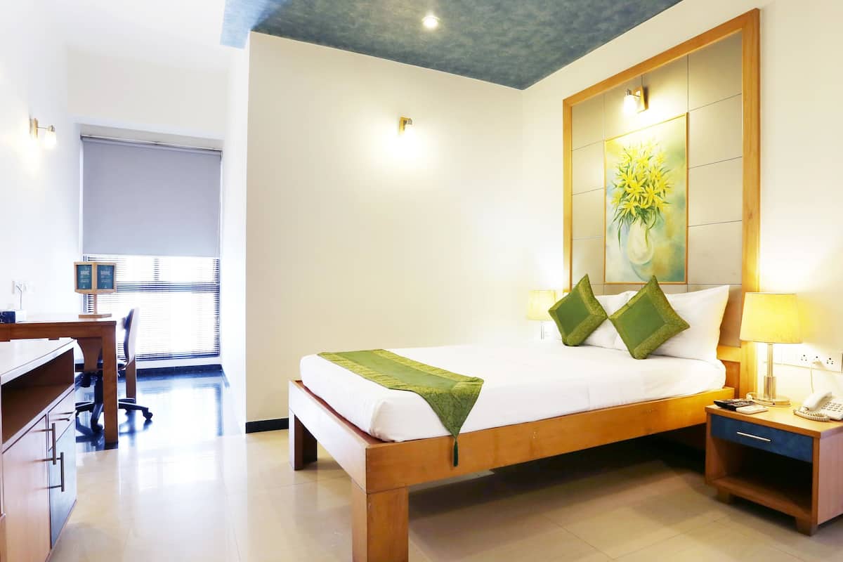 Habitación de hotel con cama, mesita de noche, lámpara y escritorio con silla. Hay una ventana con persianas y un cuadro en la pared.