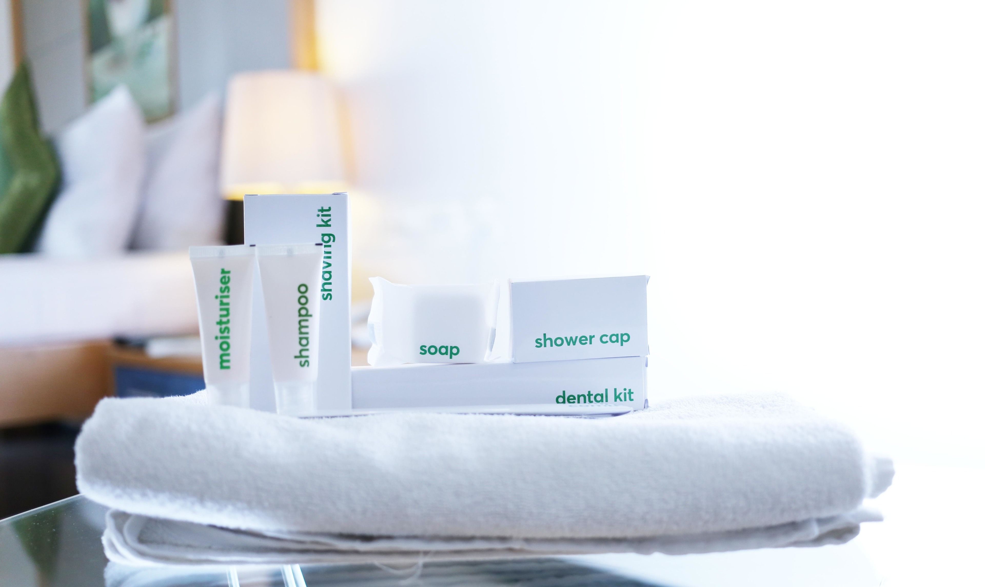 Ein ordentlich arrangiertes Set an Pflegeprodukten auf einem weißen Handtuch, darunter Zahnbürste, Zahnpasta, Seife, Shampoo, Rasierkit und eine Duschhaube.