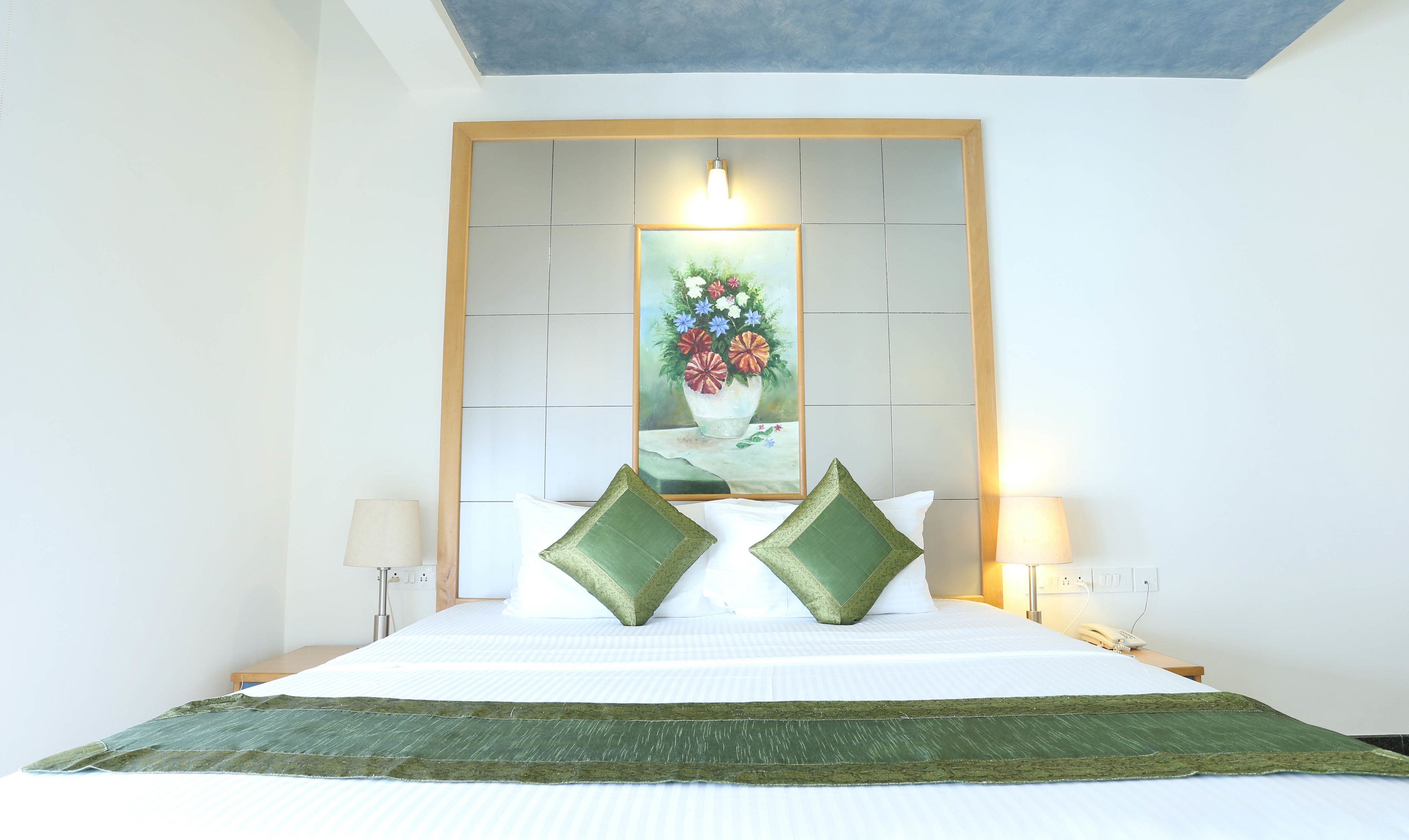 A hotel room with a large bed, two green pillows, a painting of flowers on the headboard, and bedside lamps.