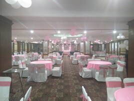 Un salón de banquetes con mesas redondas cubiertas con manteles rosas, sillas blancas adornadas con cintas rosas y un suelo alfombrado.