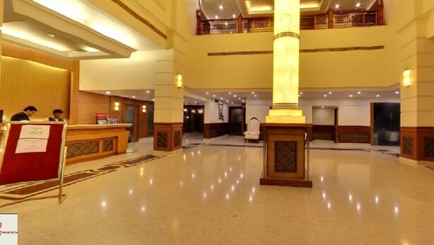A spacious hotel lobby with a central column, a reception desk, and a staircase leading to an upper level.