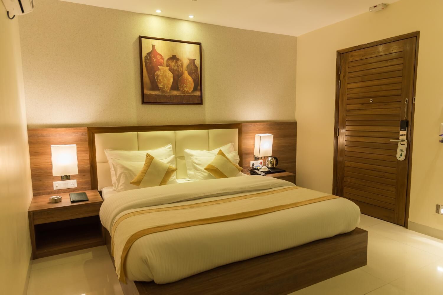 A hotel room with a large bed, two bedside tables with lamps, a wooden headboard, a painting above the bed, and a wooden door.