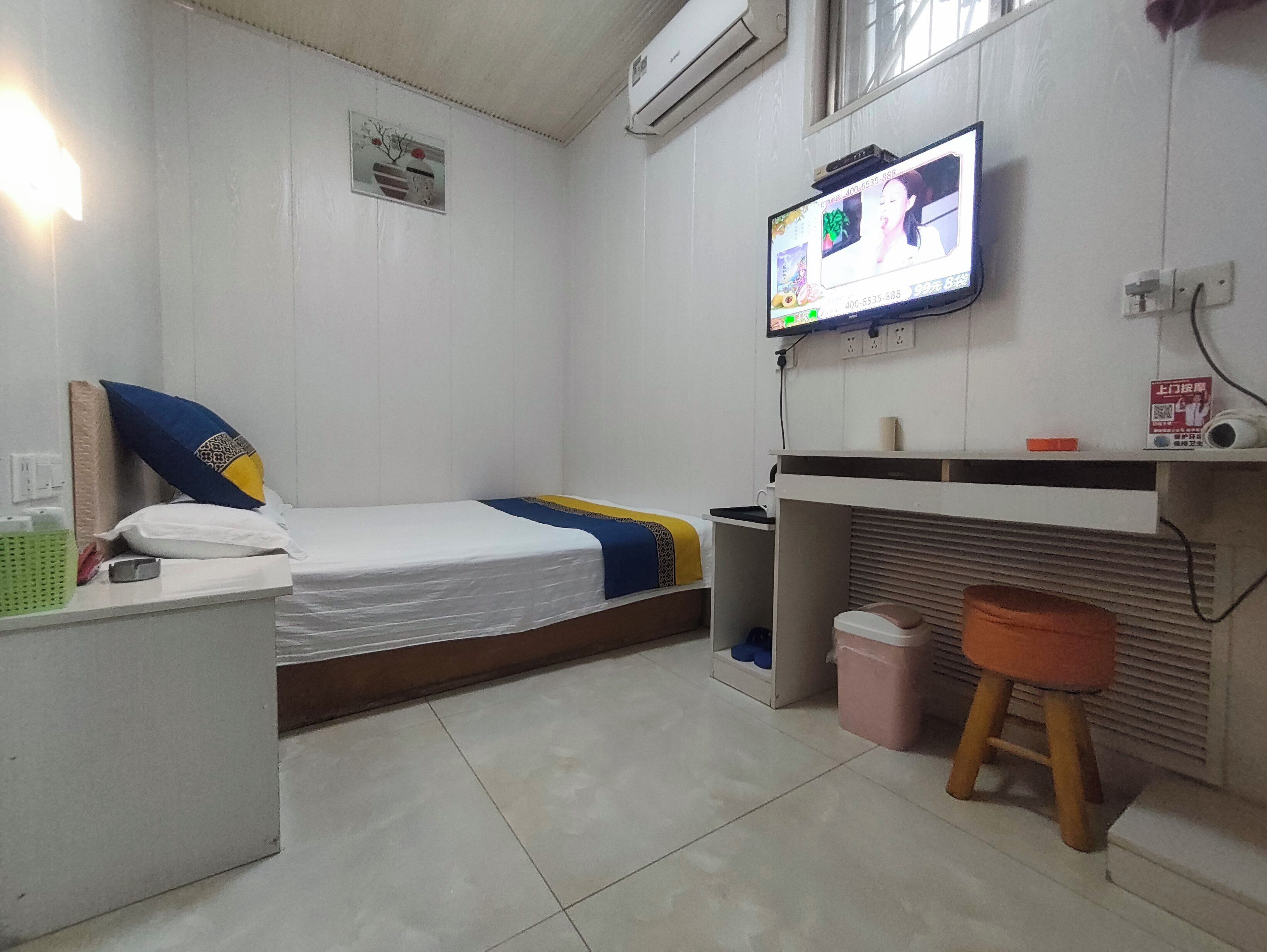 A small, clean room with a bed, a desk, a TV, and a trash bin.