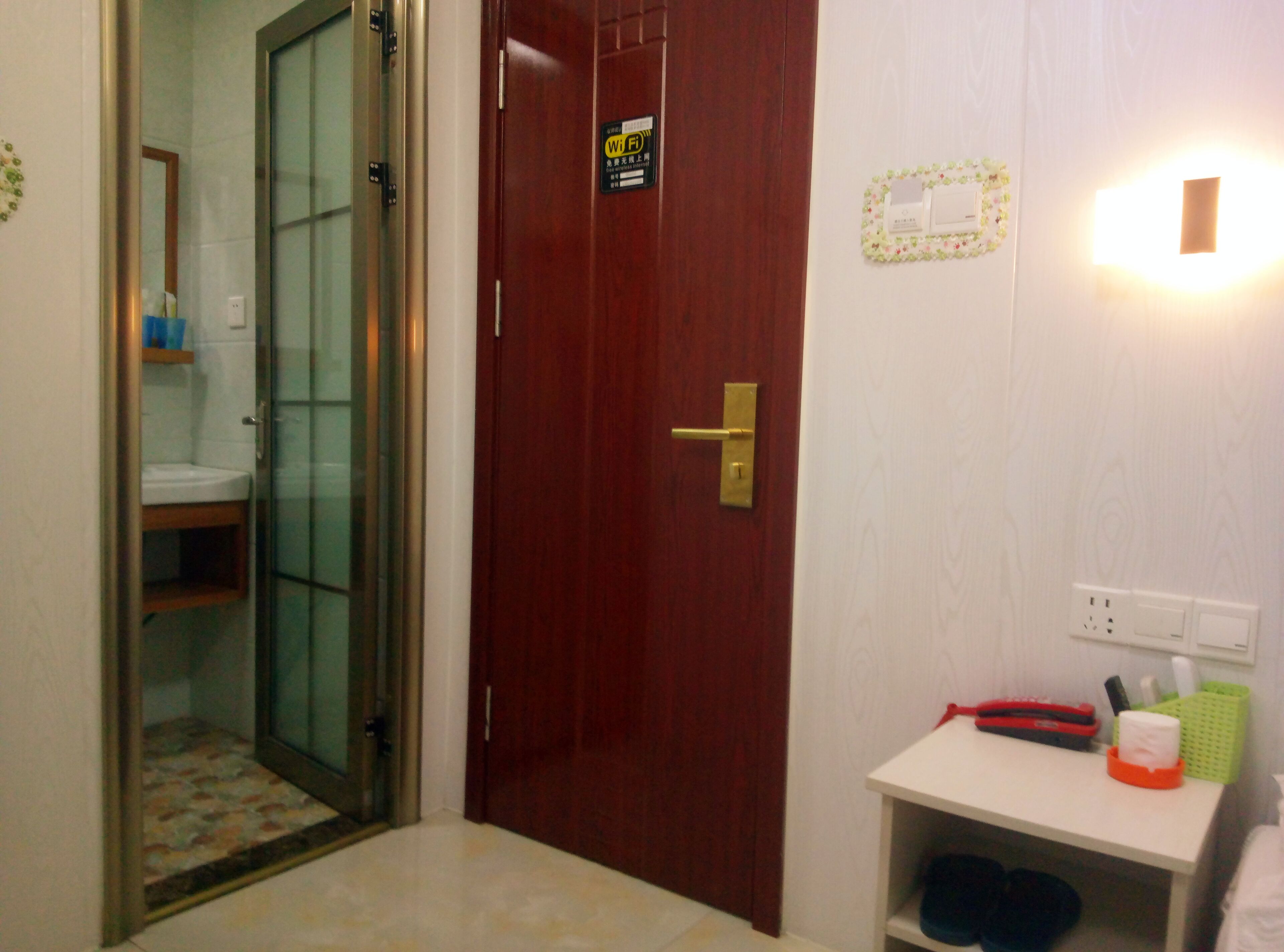Una habitación de hotel con una puerta de madera, una mesita con un objeto rojo y verde, y una lámpara fijada a la pared.