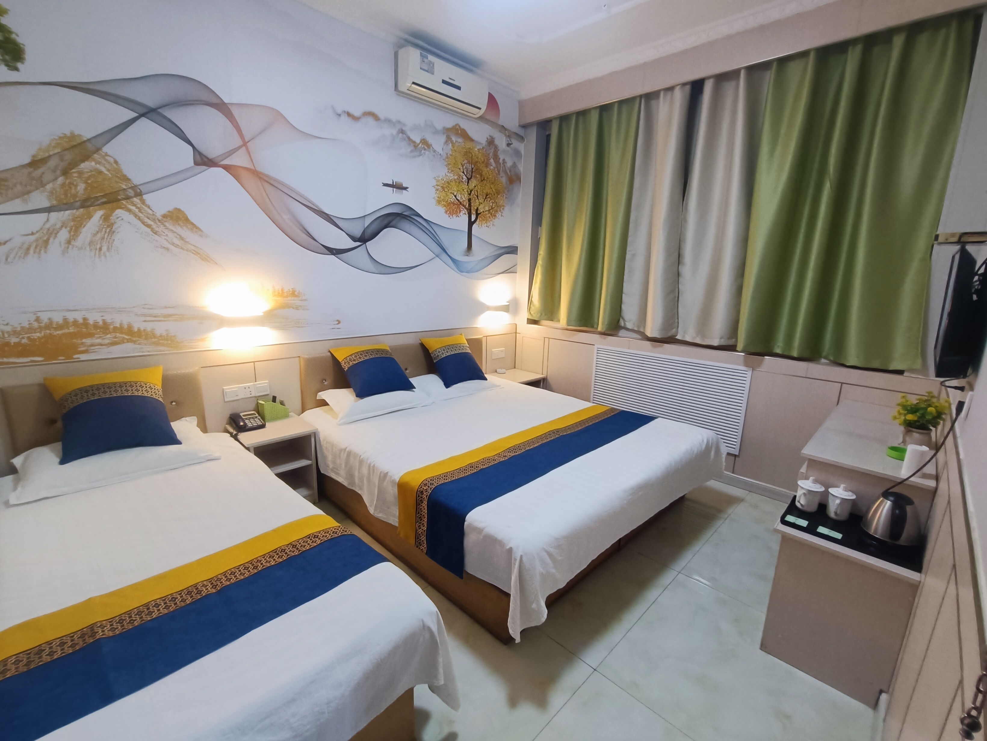 A hotel room with two beds, a mural on the wall, a kitchenette, and green curtains.