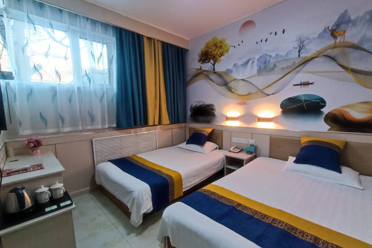 Quarto de hotel com duas camas, uma pequena mesa de trabalho com chaleira e chávenas, uma mesinha de cabeceira com telefone e um mural na parede.