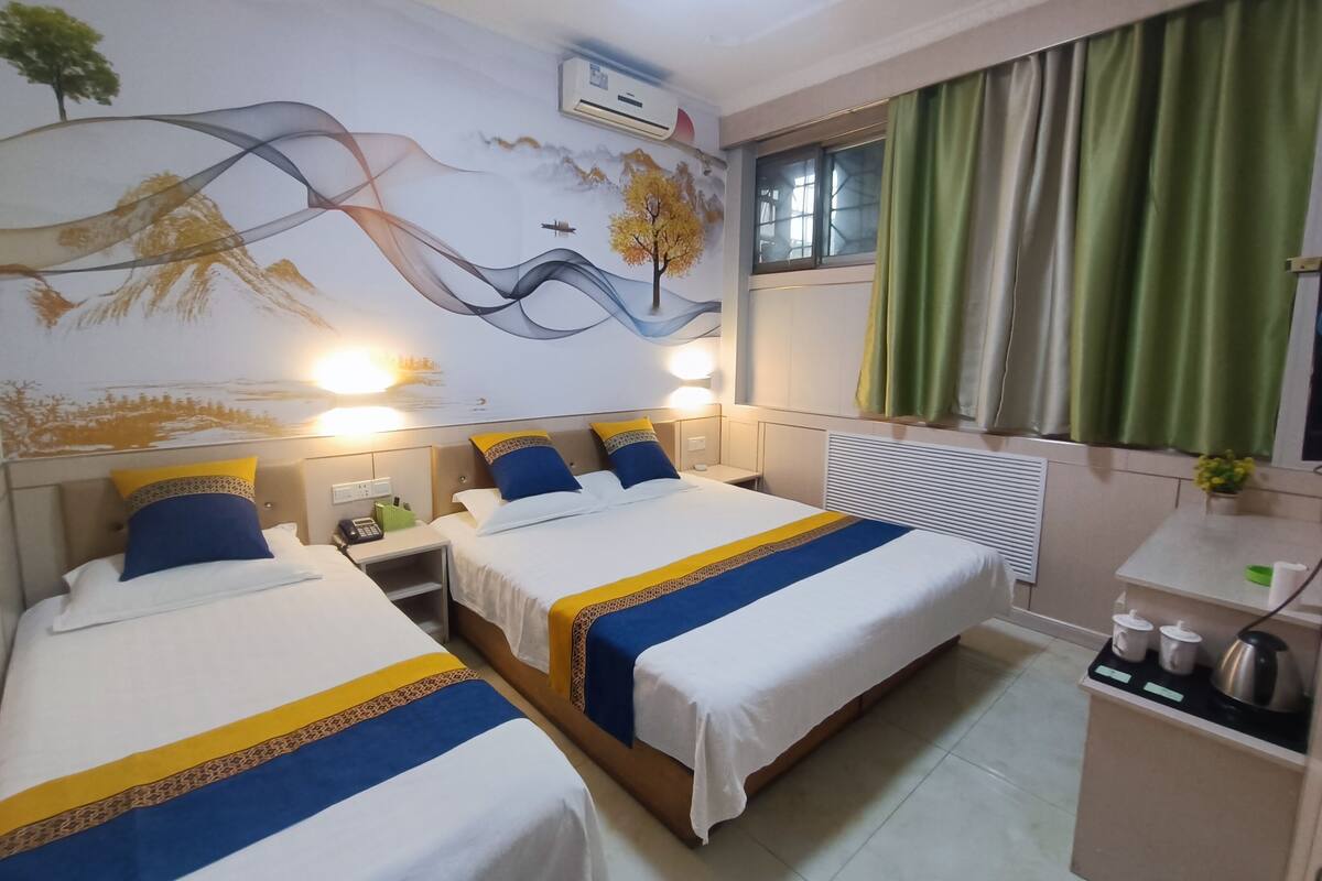 Quarto de hotel com duas camas, um mural na parede, uma janela com cortinas e uma kitchenette pequena.