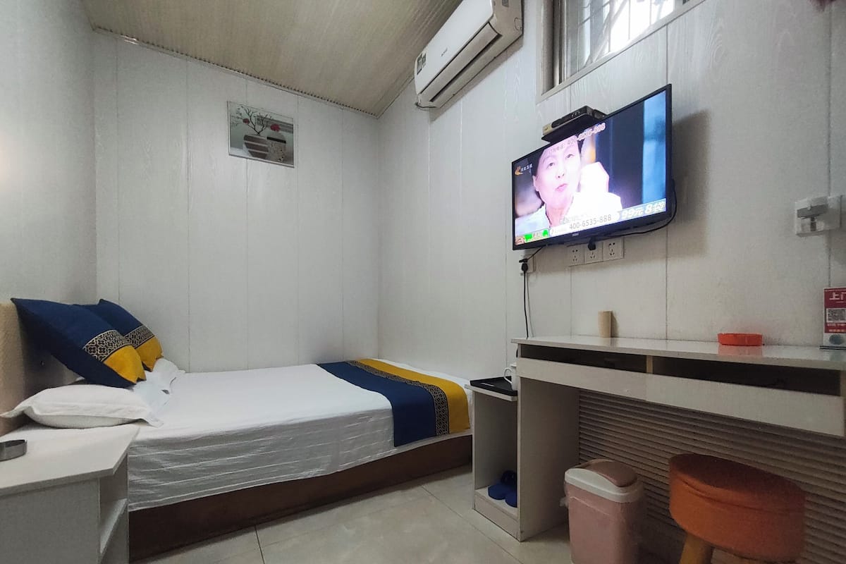 Quarto pequeno e limpo, com uma cama, escrivaninha, televisão e lixeira.
