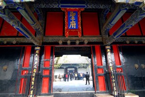 Un portón chino tradicional con intrincadas decoraciones en azul y rojo, que da acceso a un patio con un grupo de personas.