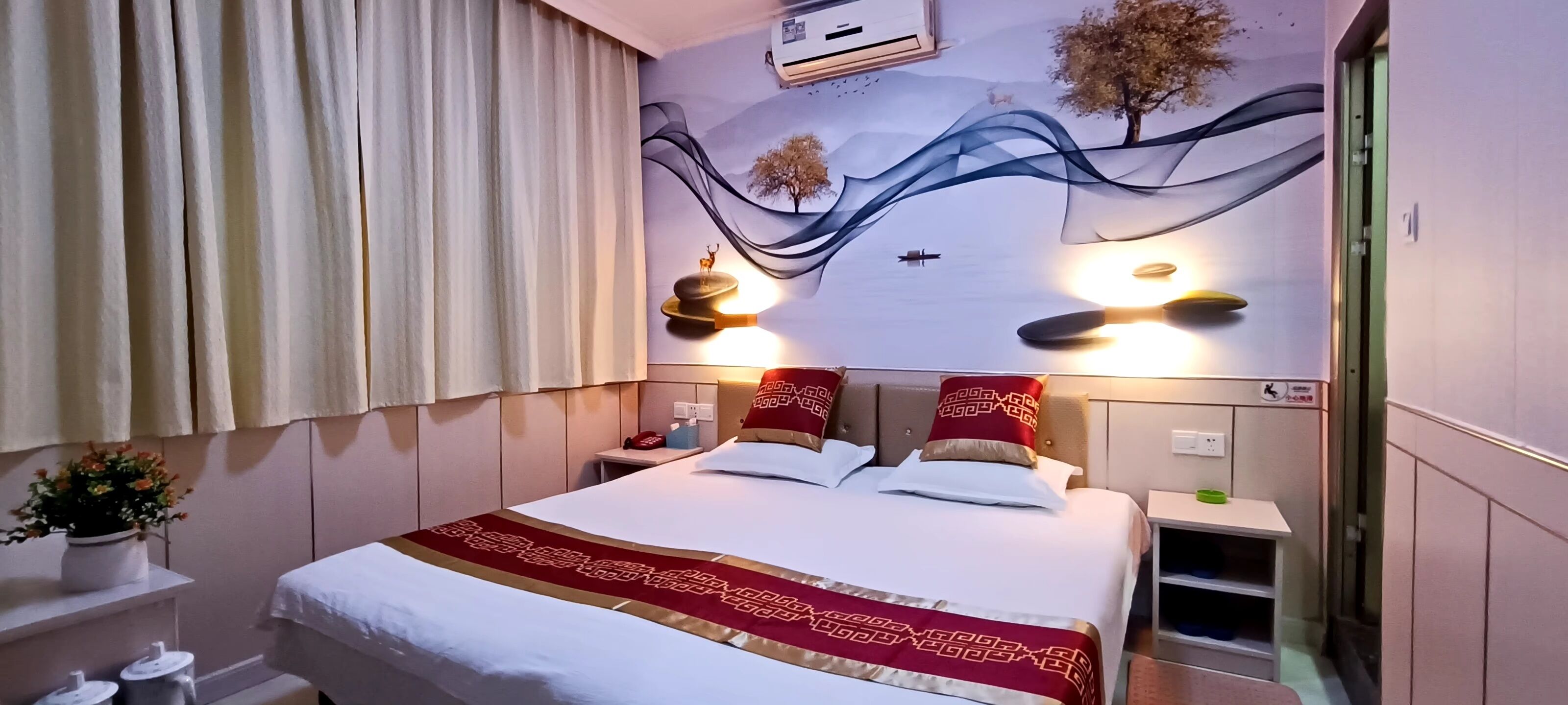 A hotel room with a bed, a nightstand, a bedside table, a wall-mounted lamp, and a window with curtains.