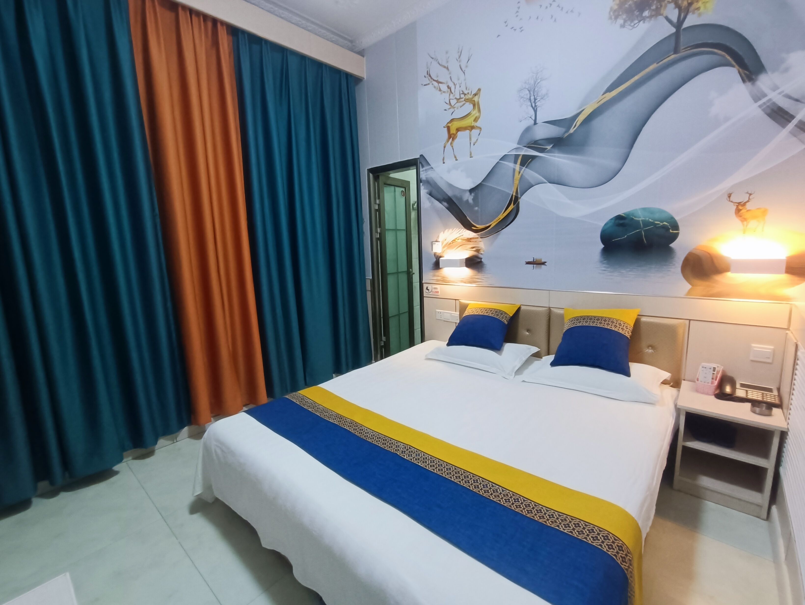 A hotel room with a bed, curtains, a wall mural, and a bedside table with a lamp.