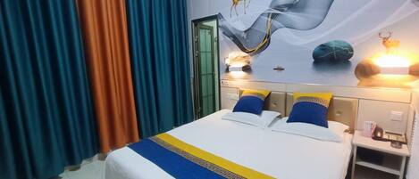 Habitación de hotel con cama, cortinas, un mural en la pared y una mesita de noche con lámpara.
