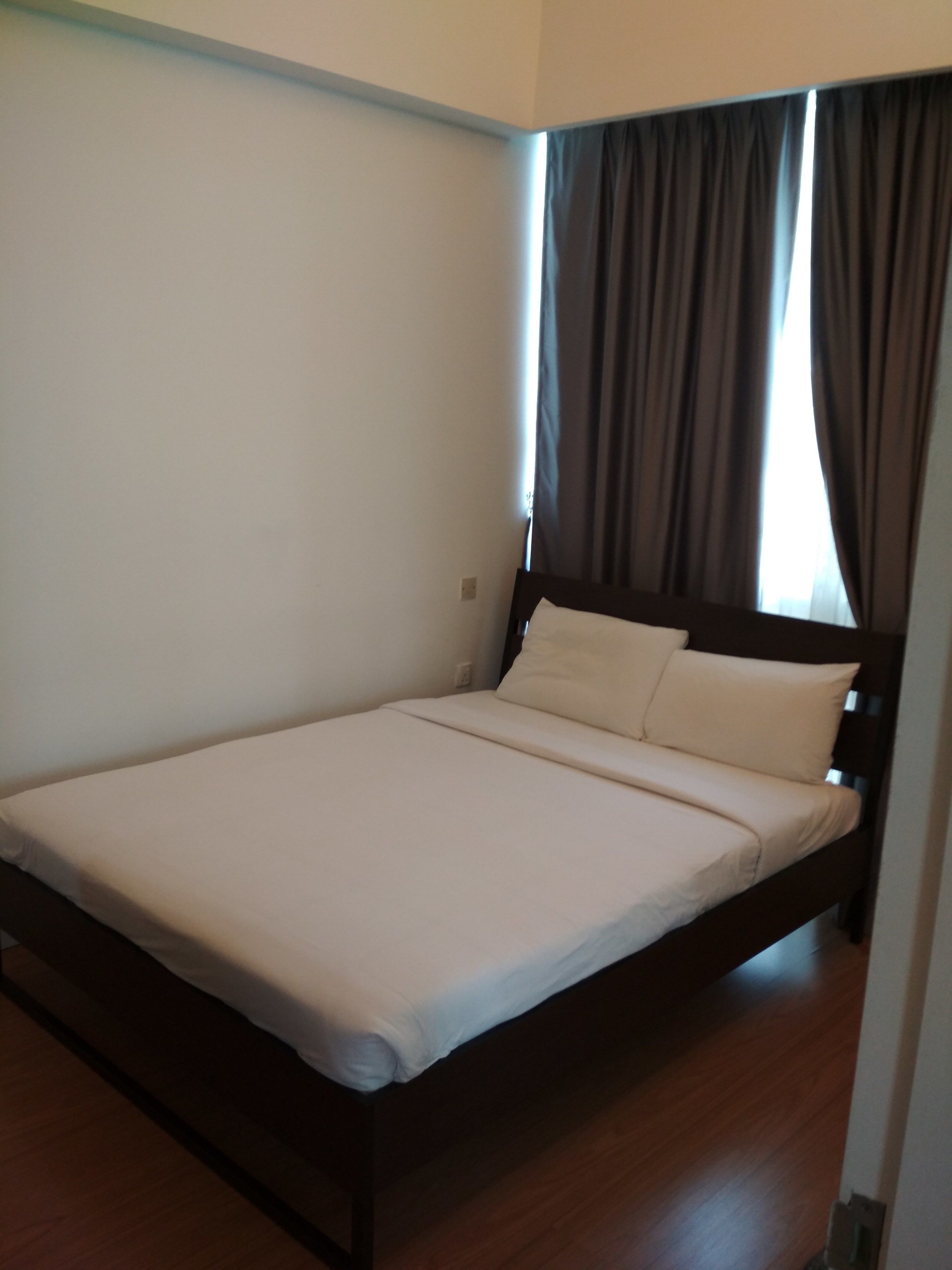 Una cama individual con estructura de madera y ropa de cama blanca en una habitación con piso de madera y una ventana con cortinas oscuras.