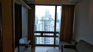 A modern living room with a large window, a sofa, a small round table, and a flat-screen TV on a stand. - Queens Service Suite at Swiss Garden Residences (Kuala Lumpur)