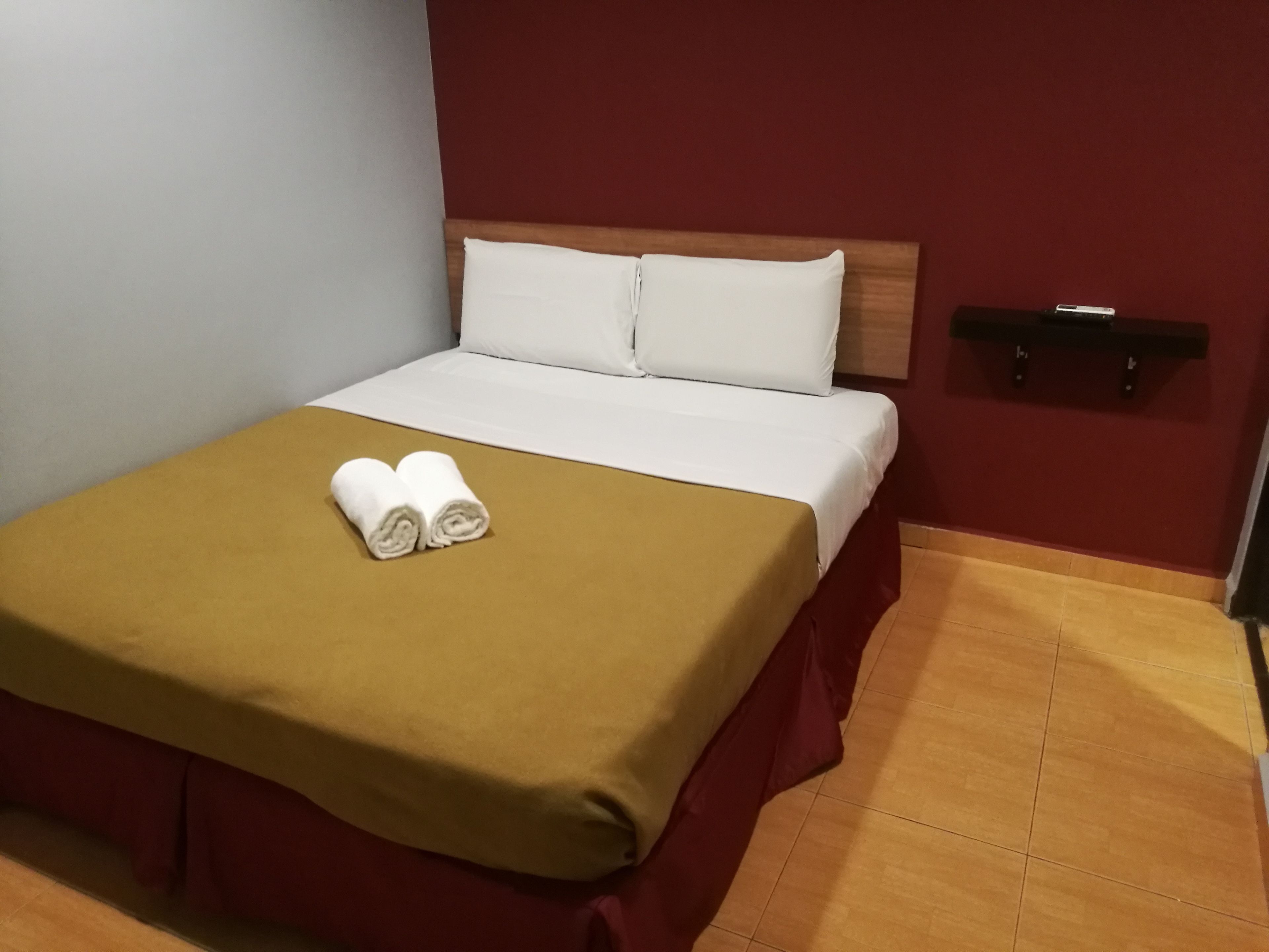 Una cama bien tendida con una colcha marrón y dos toallas enrolladas, un cabecero de madera y una pared roja con un estante negro.