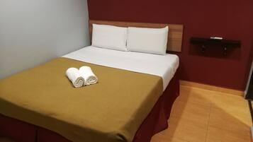 Uma cama bem arrumada com um espalha dourado e duas toalhas enroladas, uma cabeceira de madeira e uma parede vermelha com uma prateleira preta.