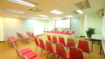 Una sala de conferencias con filas de sillas rojas, una mesa larga, una pantalla de proyector y una planta.
