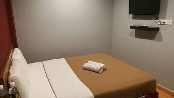 A single bed with a brown mattress and white bedding, a TV mounted on the wall, and a small shelf below it.