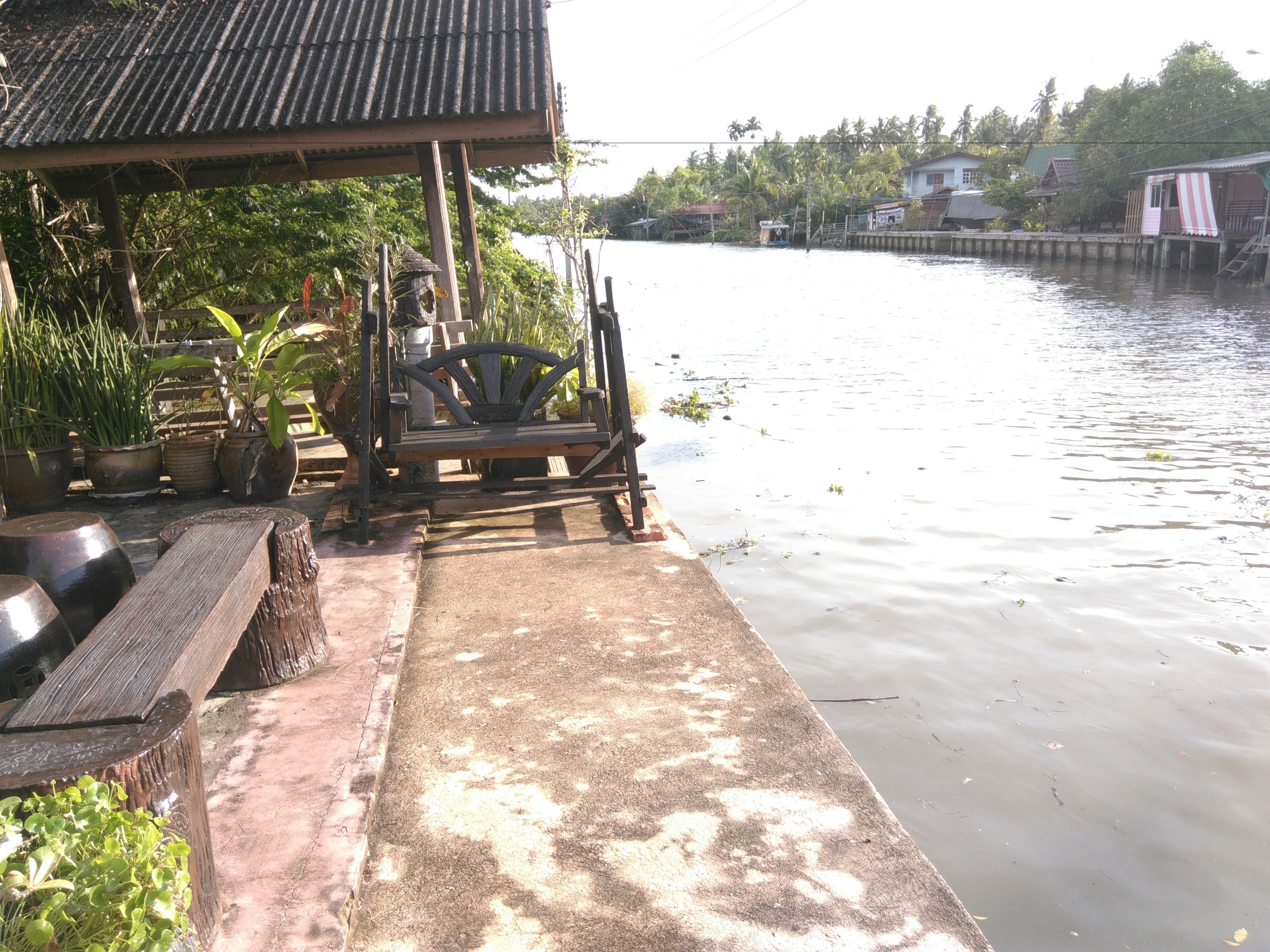 A riverside wooden bench with potted plants, a concrete pathway, and a traditional house with a tiled roof.