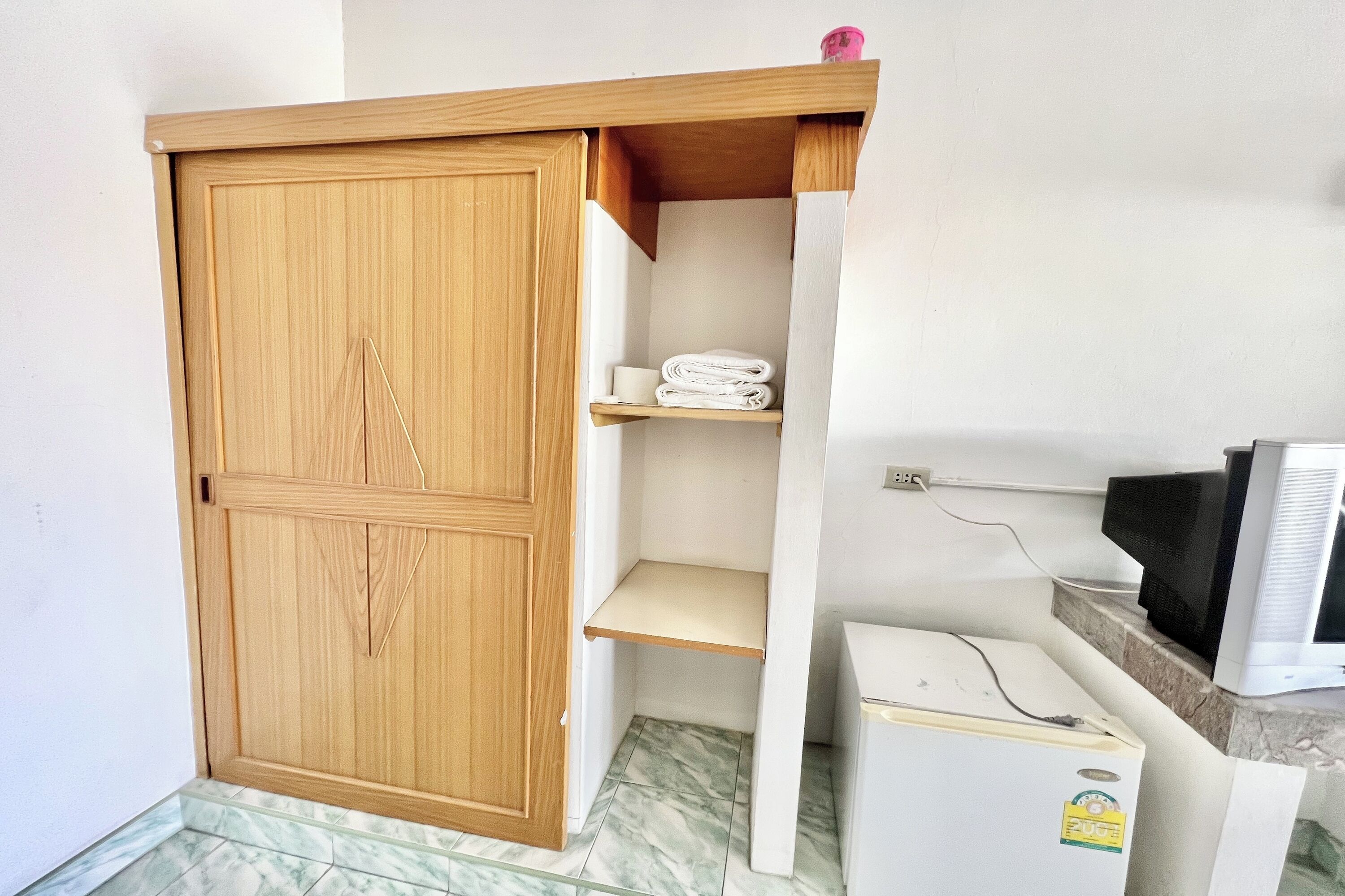 Una habitación con un armario de madera, un pequeño estante con toallas, un refrigerador blanco y un televisor sobre un piso de baldosas de mármol.