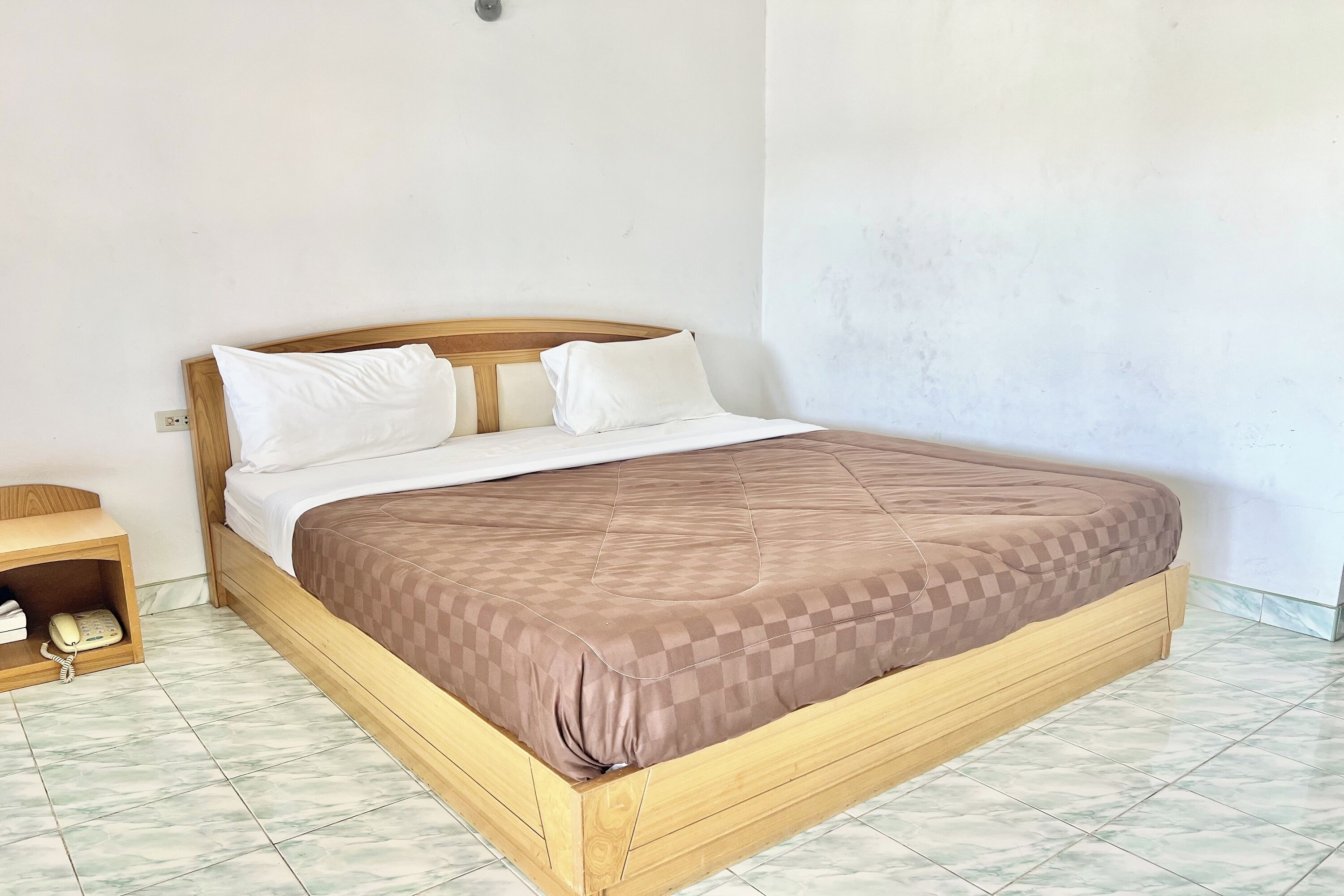 Una cama individual con estructura de madera y una colcha marrón a cuadros, una mesita de noche con un teléfono, y un aplique de pared.