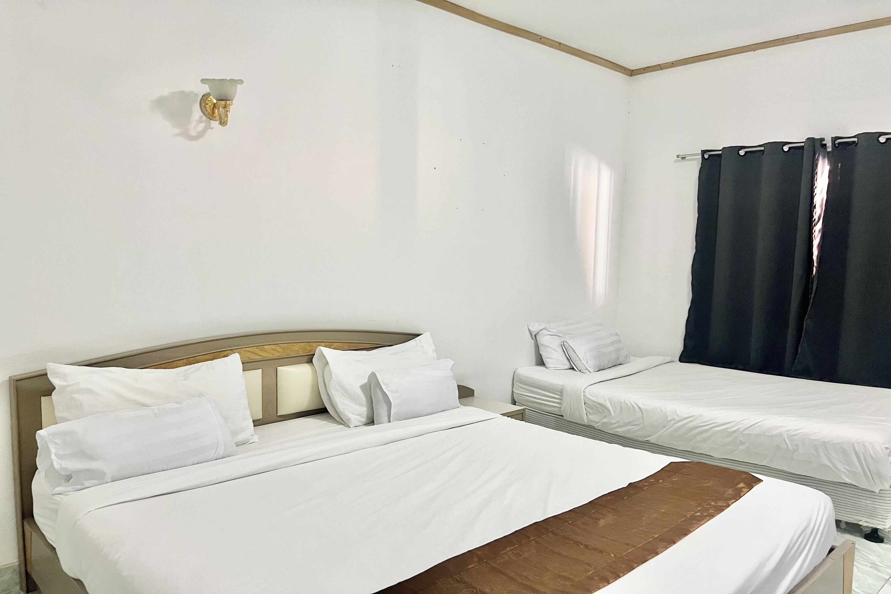 A hotel room with two single beds, a nightstand, a wall-mounted light, and a window with black curtains.